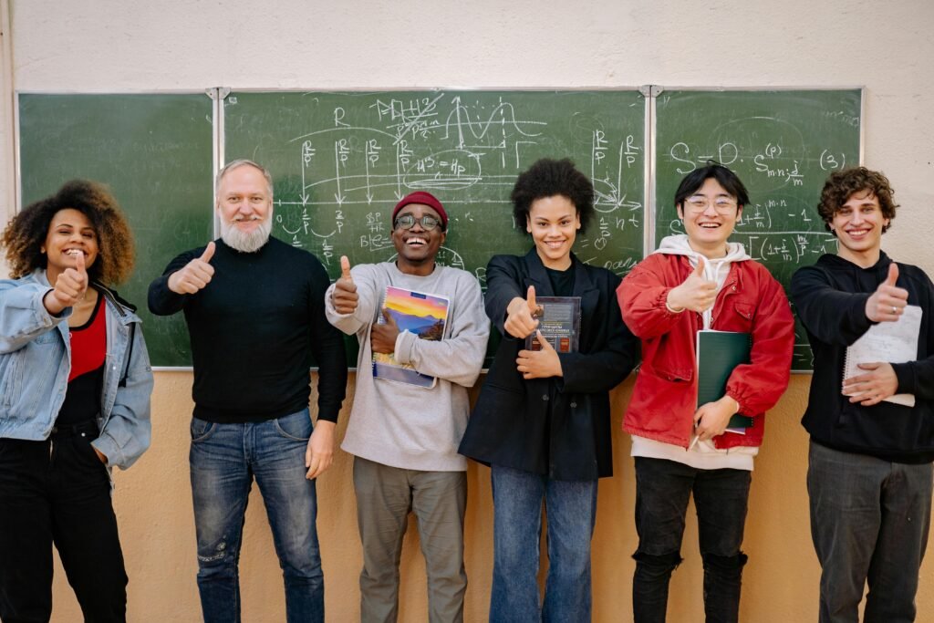 A group of diverse students with a teacher, all thumbs up, in front of a chalkboard with equations.